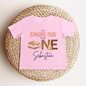 K&ouml;nnte beinhalten: Ein rosa Kleinkind-T-Shirt mit dem Text "IT'S S'MORE FUN TO BE ONE" in Orange und Braun. Eine Grafik eines S'more und eines Lagerfeuers sind ebenfalls abgebildet. Der Name "Sebastian" steht darunter in Schreibschrift.