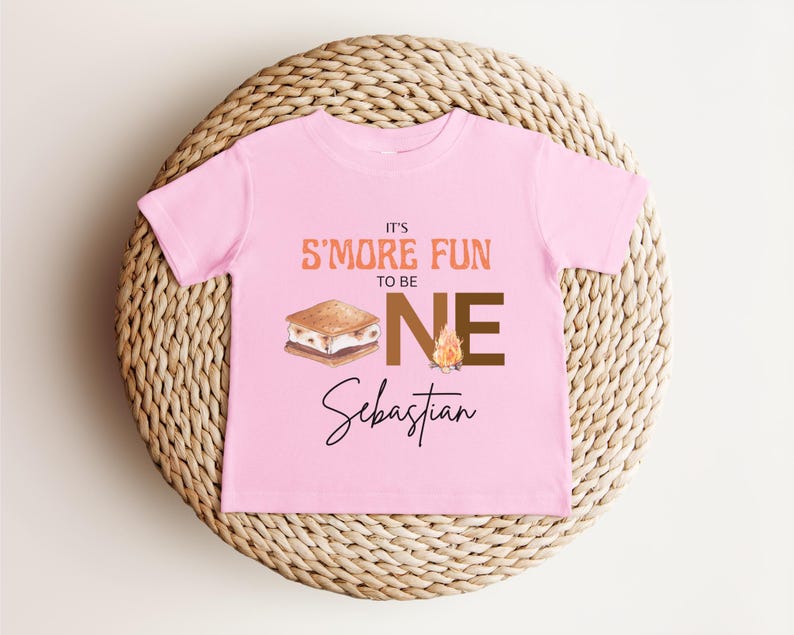 K&ouml;nnte beinhalten: Ein rosa Kleinkind-T-Shirt mit dem Text "IT'S S'MORE FUN TO BE ONE" in Orange und Braun. Eine Grafik eines S'more und eines Lagerfeuers sind ebenfalls abgebildet. Der Name "Sebastian" steht darunter in Schreibschrift.
