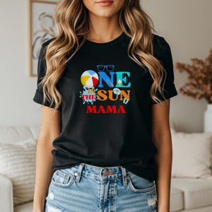 Puede incluir: Una camiseta negra de manga corta con un estampado gr&aacute;fico que incluye el texto ONE IN THE SUN MAMA. El dise&ntilde;o en el pecho muestra una pelota de playa, gafas de sol azules, un cono de helado, un salvavidas y chanclas en colores vibrantes.