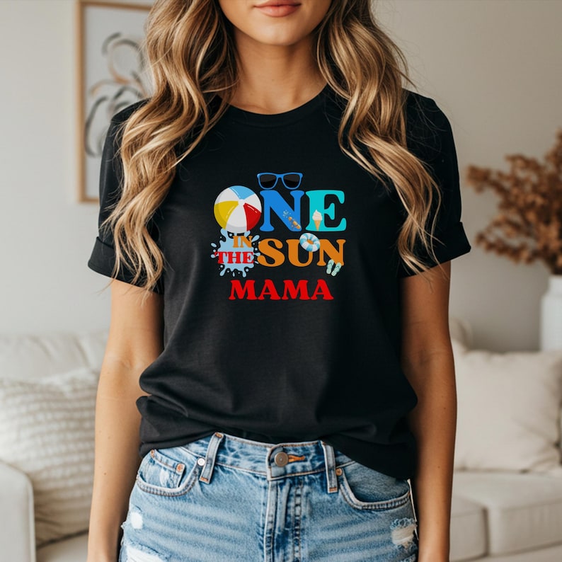 Puede incluir: Una camiseta negra de manga corta con un estampado gr&aacute;fico que incluye el texto ONE IN THE SUN MAMA. El dise&ntilde;o en el pecho muestra una pelota de playa, gafas de sol azules, un cono de helado, un salvavidas y chanclas en colores vibrantes.