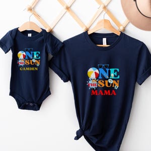 Puede incluir: Un body de manga corta azul marino y una camiseta azul marino a juego. Ambas prendas presentan un gr&aacute;fico con una pelota de playa, gafas de sol y el texto "ONE IN THE SUN". El body incluye el nombre "CAMDEN" y la camiseta "MAMA".