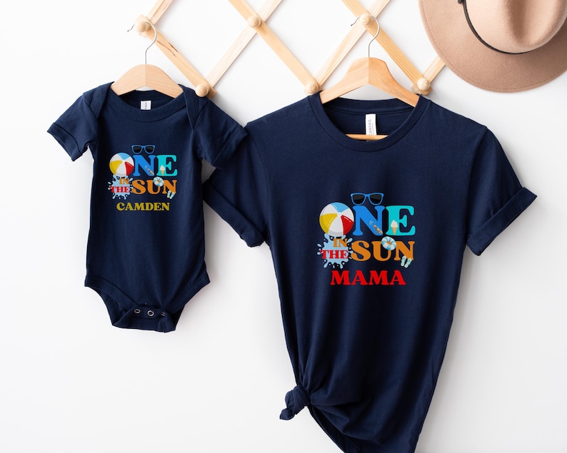 Puede incluir: Un body de manga corta azul marino y una camiseta azul marino a juego. Ambas prendas presentan un gr&aacute;fico con una pelota de playa, gafas de sol y el texto "ONE IN THE SUN". El body incluye el nombre "CAMDEN" y la camiseta "MAMA".