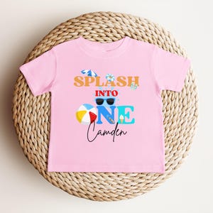 May include: A pink t-shirt with the text "Splash into One Camden" in orange, blue, and black. The design includes sunglasses, a beach ball, an ice cream cone, and a small umbrella. The shirt is displayed on a woven wicker surface.