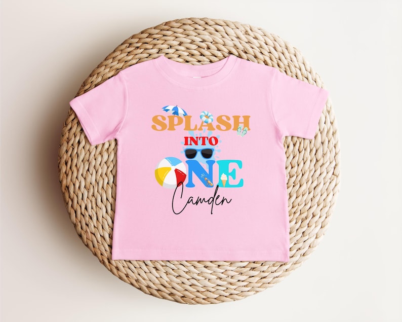 May include: A pink t-shirt with the text "Splash into One Camden" in orange, blue, and black. The design includes sunglasses, a beach ball, an ice cream cone, and a small umbrella. The shirt is displayed on a woven wicker surface.