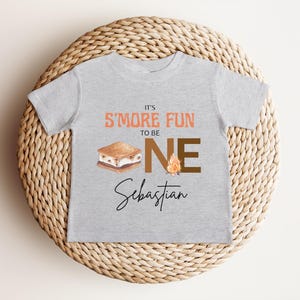 K&ouml;nnte beinhalten: Ein hellgraues T-Shirt mit dem Text "IT'S S'MORE FUN TO BE ONE" in Orange und Braun. Unter dem Text befindet sich eine Grafik eines S'more und eines Lagerfeuers. Der Name "Sebastian" ist darunter in Kursivschrift geschrieben.