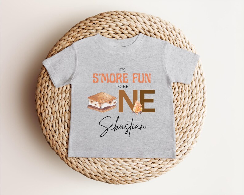 K&ouml;nnte beinhalten: Ein hellgraues T-Shirt mit dem Text "IT'S S'MORE FUN TO BE ONE" in Orange und Braun. Unter dem Text befindet sich eine Grafik eines S'more und eines Lagerfeuers. Der Name "Sebastian" ist darunter in Kursivschrift geschrieben.