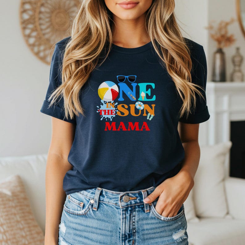 Puede incluir: Una camiseta azul marino de manga corta con un estampado gr&aacute;fico colorido que dice ONE IN THE SUN MAMA. El dise&ntilde;o incluye una pelota de playa, gafas de sol, un helado, un flotador y chanclas en tonos brillantes de azul, amarillo y rojo.