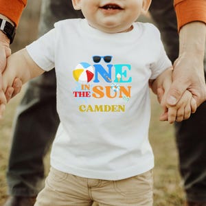 Puede incluir: Una camiseta blanca de manga corta con un gr&aacute;fico colorido que incluye el texto ONE IN THE SUN y el nombre CAMDEN impreso debajo. El dise&ntilde;o presenta una pelota de playa, gafas de sol, un cono de helado y un salvavidas en azul, naranja y amarillo.