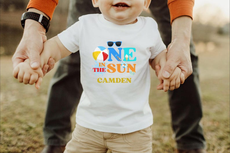 Puede incluir: Una camiseta blanca de manga corta con un gr&aacute;fico colorido que incluye el texto ONE IN THE SUN y el nombre CAMDEN impreso debajo. El dise&ntilde;o presenta una pelota de playa, gafas de sol, un cono de helado y un salvavidas en azul, naranja y amarillo.