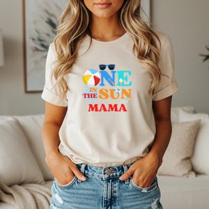 Puede incluir: Una camiseta beige de manga corta con un estampado gr&aacute;fico que incluye el texto "ONE IN THE SUN MAMA". El dise&ntilde;o presenta una pelota de playa colorida, gafas de sol negras, un peque&ntilde;o cono de helado, un salvavidas y chanclas en azul, naranja y rojo.
