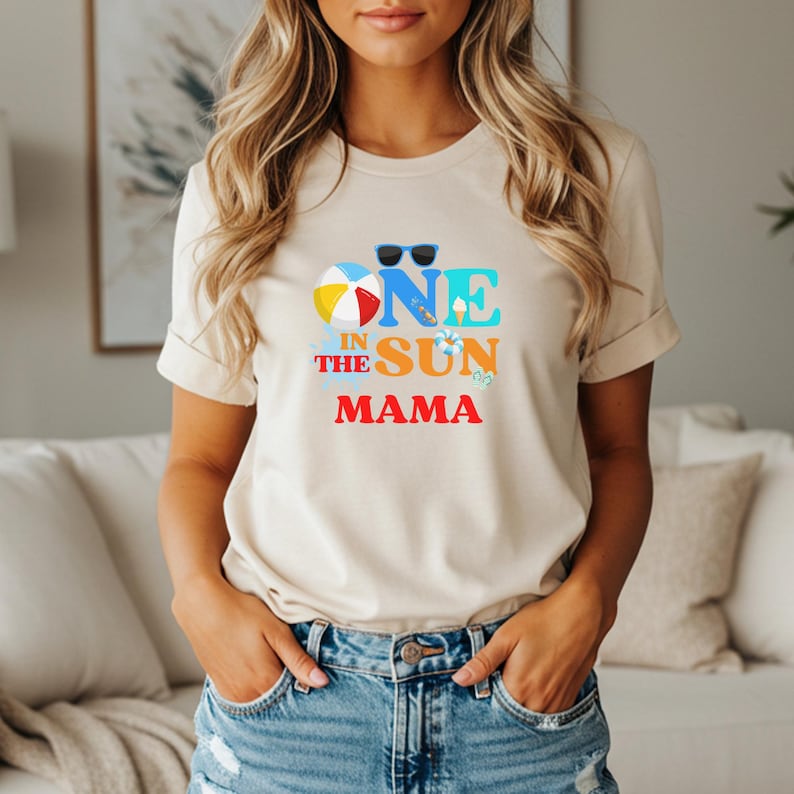 Puede incluir: Una camiseta beige de manga corta con un estampado gr&aacute;fico que incluye el texto "ONE IN THE SUN MAMA". El dise&ntilde;o presenta una pelota de playa colorida, gafas de sol negras, un peque&ntilde;o cono de helado, un salvavidas y chanclas en azul, naranja y rojo.