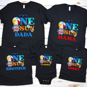 Puede incluir: Un conjunto de cinco camisetas negras y un body negro con un estampado gr&aacute;fico con el texto ONE IN THE SUN. El dise&ntilde;o incluye una pelota de playa, gafas de sol e iconos de helados. Las camisetas est&aacute;n etiquetadas como DADA, MAMA, BROTHER, CAMDEN y SISSY.