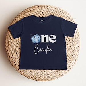 May include: A navy blue toddler's t-shirt featuring the word "one" in white, a blue watercolour pumpkin illustration, and the name "Camden". The t-shirt is displayed on a woven, circular mat.