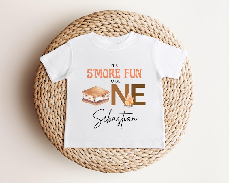 May include: White toddler t-shirt with the text "It's S'more Fun to be ONE" in orange and brown. A graphic of a s'more and a campfire are also on the shirt. The name "Sebastian" is written in cursive below the text.