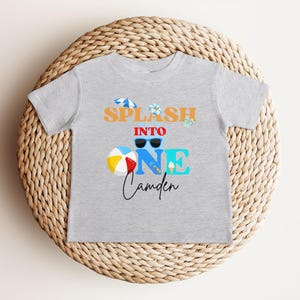 May include: A light gray t-shirt with the words "SPLASH INTO ONE Camden" in orange, blue, and black. The design includes sunglasses, a beach ball, and an ice cream cone. The shirt is displayed on a woven, circular surface.