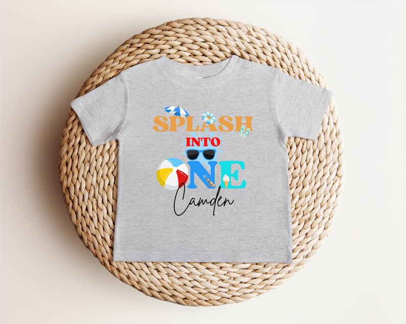 May include: A light gray t-shirt with the words "SPLASH INTO ONE Camden" in orange, blue, and black. The design includes sunglasses, a beach ball, and an ice cream cone. The shirt is displayed on a woven, circular surface.
