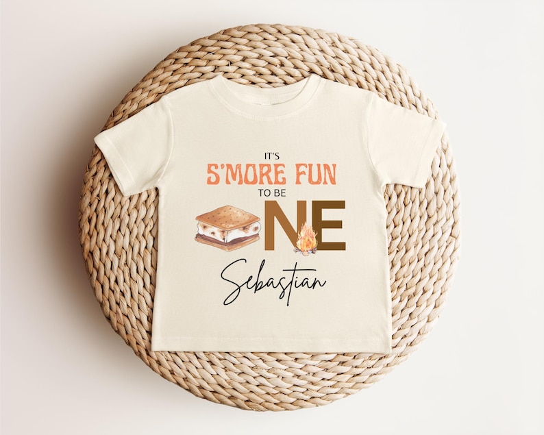 K&ouml;nnte beinhalten: Ein cremefarbenes Kleinkind-T-Shirt mit dem Text "It's S'more Fun to be ONE" in Orange und Braun. Eine Grafik eines S'more und eines Lagerfeuers sind ebenfalls abgebildet. Der Name "Sebastian" steht darunter in Schreibschrift.