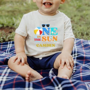Puede incluir: Una camiseta de manga corta color crema con un estampado gr&aacute;fico que dice ONE IN THE SUN, junto con una pelota de playa, gafas de sol y el nombre CAMDEN en letras amarillas. La camiseta se combina con pantalones cortos azul oscuro.