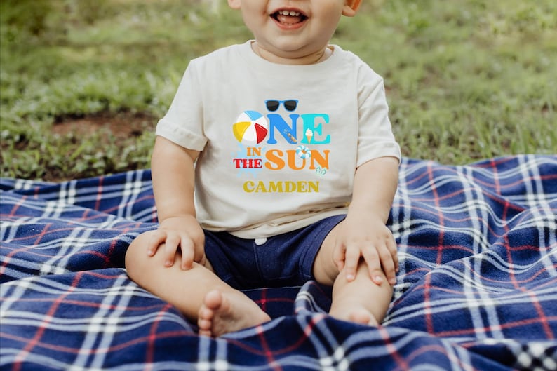 Puede incluir: Una camiseta de manga corta color crema con un estampado gr&aacute;fico que dice ONE IN THE SUN, junto con una pelota de playa, gafas de sol y el nombre CAMDEN en letras amarillas. La camiseta se combina con pantalones cortos azul oscuro.