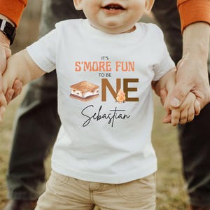 K&ouml;nnte beinhalten: Wei&szlig;es T-Shirt mit dem Text "It's S'more Fun to be ONE Sebastian". Das Design beinhaltet eine S'more-Grafik und eine Lagerfeuer-Illustration. Das T-Shirt wird von einem Erwachsenen gehalten.