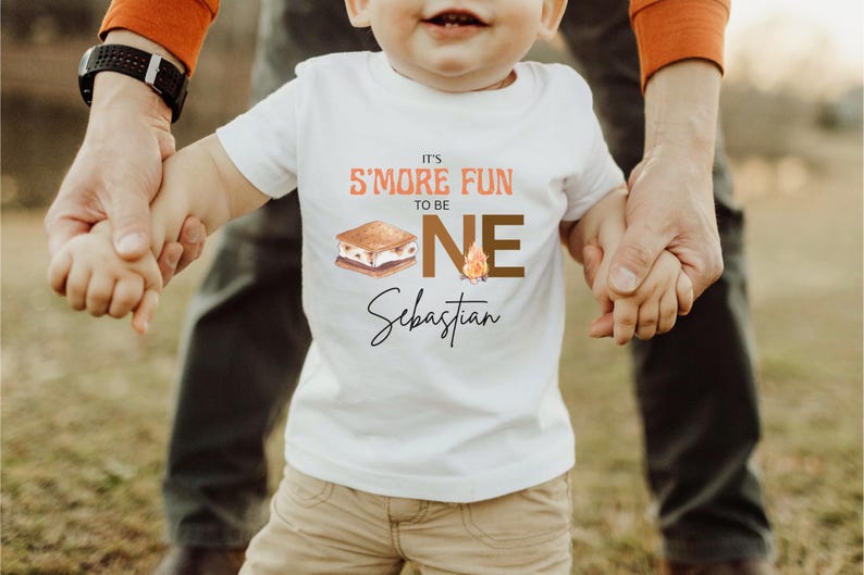 K&ouml;nnte beinhalten: Wei&szlig;es T-Shirt mit dem Text "It's S'more Fun to be ONE Sebastian". Das Design beinhaltet eine S'more-Grafik und eine Lagerfeuer-Illustration. Das T-Shirt wird von einem Erwachsenen gehalten.