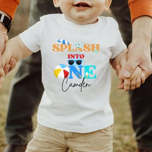 May include: White t-shirt with a colorful graphic that reads "Splash into One Camden." The design includes sunglasses, a beach ball, and an ice cream cone. The shirt is being held by an adult.