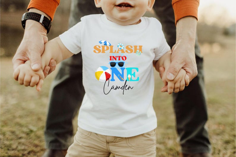 May include: White t-shirt with a colorful graphic that reads "Splash into One Camden." The design includes sunglasses, a beach ball, and an ice cream cone. The shirt is being held by an adult.
