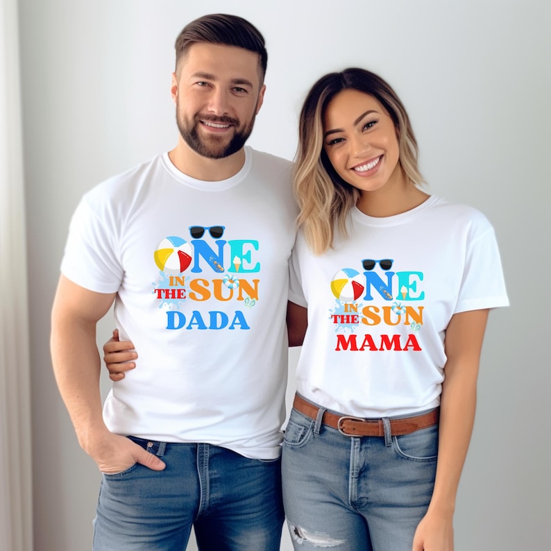May include: Two white t-shirts with the text "ONE IN THE SUN DADA" and "ONE IN THE SUN MAMA" in blue, yellow, and red. The shirts feature a beach ball and sunglasses graphic. The people are wearing blue jeans.