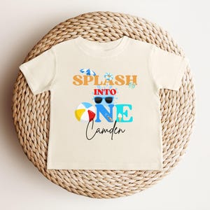 May include: Cream-colored toddler t-shirt with the text "Splash into One Camden" in orange, blue, and black. The design includes sunglasses, a beach ball, and an ice cream cone. The shirt is displayed on a woven wicker placemat.