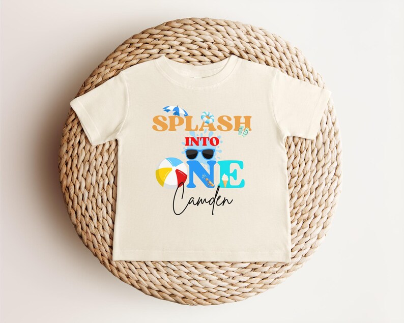 May include: Cream-colored toddler t-shirt with the text "Splash into One Camden" in orange, blue, and black. The design includes sunglasses, a beach ball, and an ice cream cone. The shirt is displayed on a woven wicker placemat.