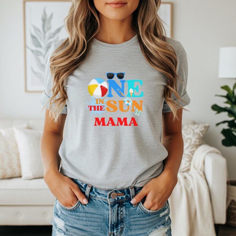 Puede incluir: Una camiseta gris jaspeado de manga corta con un estampado gr&aacute;fico que incluye el texto "ONE IN THE SUN MAMA" en azul, naranja y rojo. El dise&ntilde;o presenta iconos de una pelota de playa, gafas de sol, un helado, un salvavidas y chanclas.