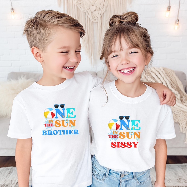 Puede incluir: Dos camisetas blancas de manga corta con un estampado gr&aacute;fico colorido. El dise&ntilde;o incluye una pelota de playa, gafas de sol y el texto ONE IN THE SUN BROTHER en una camiseta y ONE IN THE SUN SISSY en la otra, con salpicaduras de agua azul.