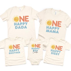 Könnte beinhalten: Eine Sammlung von cremefarbenen T-Shirts und einem Body. Jedes Kleidungsstück zeigt eine lächelnde Sonne und die Wörter "ONE" und "HAPPY" in Orange und Blau, mit Familientiteln wie "DADA", "MAMA", "BROTHER" und "SISSY". Der Body sagt "HAPPY BOY Camden".