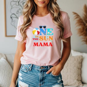 Puede incluir: Una camiseta de manga corta de color rosa claro con un estampado gr&aacute;fico que incluye el texto ONE IN THE SUN MAMA en azul, naranja y rojo. El dise&ntilde;o presenta ilustraciones de gafas de sol azules, una pelota de playa, un helado y chanclas.
