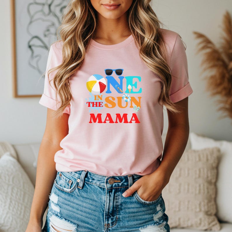 Puede incluir: Una camiseta de manga corta de color rosa claro con un estampado gr&aacute;fico que incluye el texto ONE IN THE SUN MAMA en azul, naranja y rojo. El dise&ntilde;o presenta ilustraciones de gafas de sol azules, una pelota de playa, un helado y chanclas.