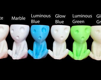 May include: Seven small, white ceramic figurines with different colored glazes. The figurines are sitting down with their hands on their laps. The glazes are white, marble, luminous blue, glow blue, luminous green, and glow green.