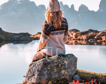 Könnte beinhalten: Eine Frau in einem beigefarbenen Strickpullover mit einem braunen, schwarzen und weißen geometrischen Muster sitzt auf einem großen Felsen mit Blick auf einen See. Sie trägt eine beigefarbene Strickmütze mit einem weißen Bommel. Die Berge sind im Hintergrund.