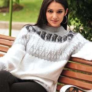 Op de afbeelding: Een vrouw draagt een witte en grijze alpaca trui met een geometrisch patroon. De trui heeft een ronde hals en lange mouwen. De vrouw zit op een houten bank.