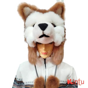 Op de afbeelding: Een pluche vossenmuts in bruin, wit en zwart, met een gedetailleerd gezicht en oren. De muts wordt gedragen over een witte zip-up trui met een patroon en bruine pompons. Het Kintu Alpaca-logo is zichtbaar.