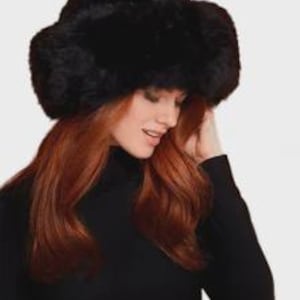 May include: A black faux fur trapper hat with a thick, plush texture and ear flaps that extend down the sides. The hat features a rounded crown and a dense, soft appearance, providing full coverage for the head and ears in a solid black colour.