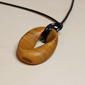 May include: A wooden pendant necklace featuring a light brown, oval-shaped pendant. The pendant is suspended from a black cord, knotted at the top. The wood grain is visible, adding texture to the natural-toned jewelry.