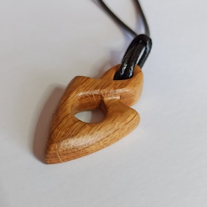Könnte beinhalten: Ein Holzanhänger in Form eines stilisierten Herzens mit einem Loch in der Mitte, der an einer schwarzen Schnur hängt.