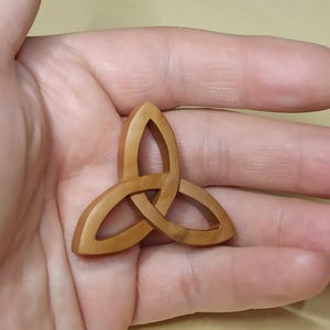 Könnte beinhalten: Ein hellbrauner Holzanhänger mit einem Triquetra-Knoten. Das Design ist glatt und poliert. Das Triquetra ist ein Symbol für Verbundenheit und wird oft in keltischer Kunst und Schmuck verwendet.