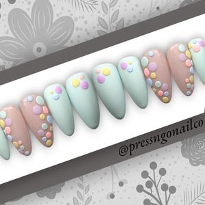 May include: A set of ten long, almond-shaped press-on nails with a pastel green and light brown base. Each nail is decorated with colorful polka dots.
