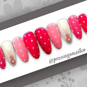 May include: Set of ten pink and red press-on nails with white polka dots. One nail features a white bunny silhouette. The nails are arranged in a row on a white surface. The text "@pressngonailco" is visible in the lower right corner of the image.