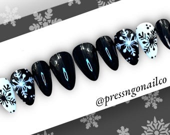 Puede incluir: Un conjunto de uñas postizas negras brillantes con un diseño de copo de nieve. Algunas uñas son negras lisas, mientras que otras presentan copos de nieve blancos sobre una base negra o azul claro. Las uñas tienen forma ovalada y están dispuestas en fila.