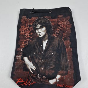 Puede incluir: Bolsa negra con cordón con un retrato en tonos sepia de un hombre con una camisa oscura sobre un diseño de pared de ladrillos. La bolsa incluye el texto "B. Tsoi" y los años "1962-1990" en rojo.