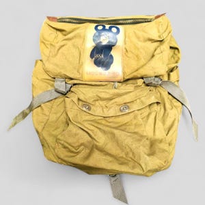 May include: A vintage olive green backpack with a zipper closure and adjustable straps. The front features a patch with a cartoon bear mascot and the text "MOCKBA-80". The backpack has two front pockets with snap closures.