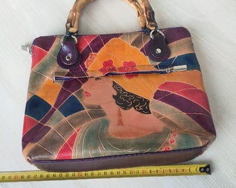 Puede incluir: Un bolso de mano de cuero pintado a mano, con un asa de bambú. El bolso presenta un diseño geométrico con un retrato de mujer. El bolso mide aproximadamente 30 cm de ancho.