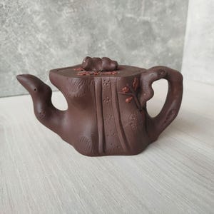 Puede incluir: Una tetera de cerámica marrón con un diseño único y orgánico. La tetera presenta un asa y un pico esculpidos, con detalles tallados y una tapa. La tetera es de color marrón oscuro y parece hecha a mano.